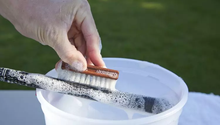 Using a brush to clean golf clubs over a bucket of soapy water